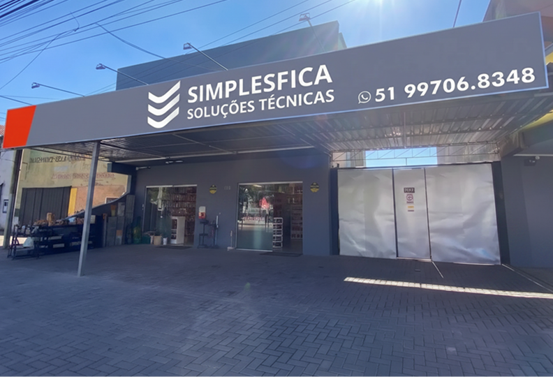Fachada externa da loja física da SIMPLESFICA Soluções Técnicas, na Avenida Rio Grande do Sul, 805, Mathias Velho, Canoas, Rio Grande do Sul. A imagem mostra a frente do estabelecimento com calçada em blocos intertravados cinza e um letreiro moderno na cor cinza-escuro com o logotipo em formato de três setas brancas e o número de atendimento em destaque: (51) 99706.8348. A loja fica aberta 24h por dia, todos os dias da semana.