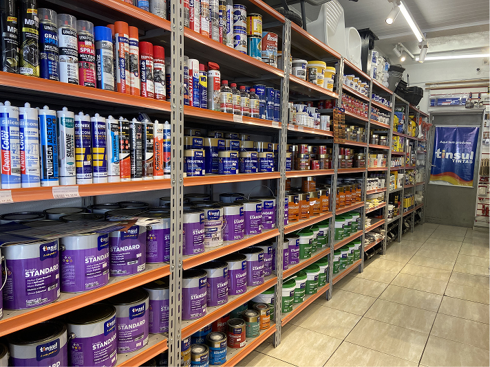 Corredor dedicado a acabamentos e produtos químicos. Estantes metálicas industriais exibem fileiras organizadas de latas de tintas imobiliárias, vernizes e impermeabilizantes em tons de roxo, azul e verde. Na prateleira superior esquerda, estão alinhados diversos tubos aplicadores de selantes e silicones.